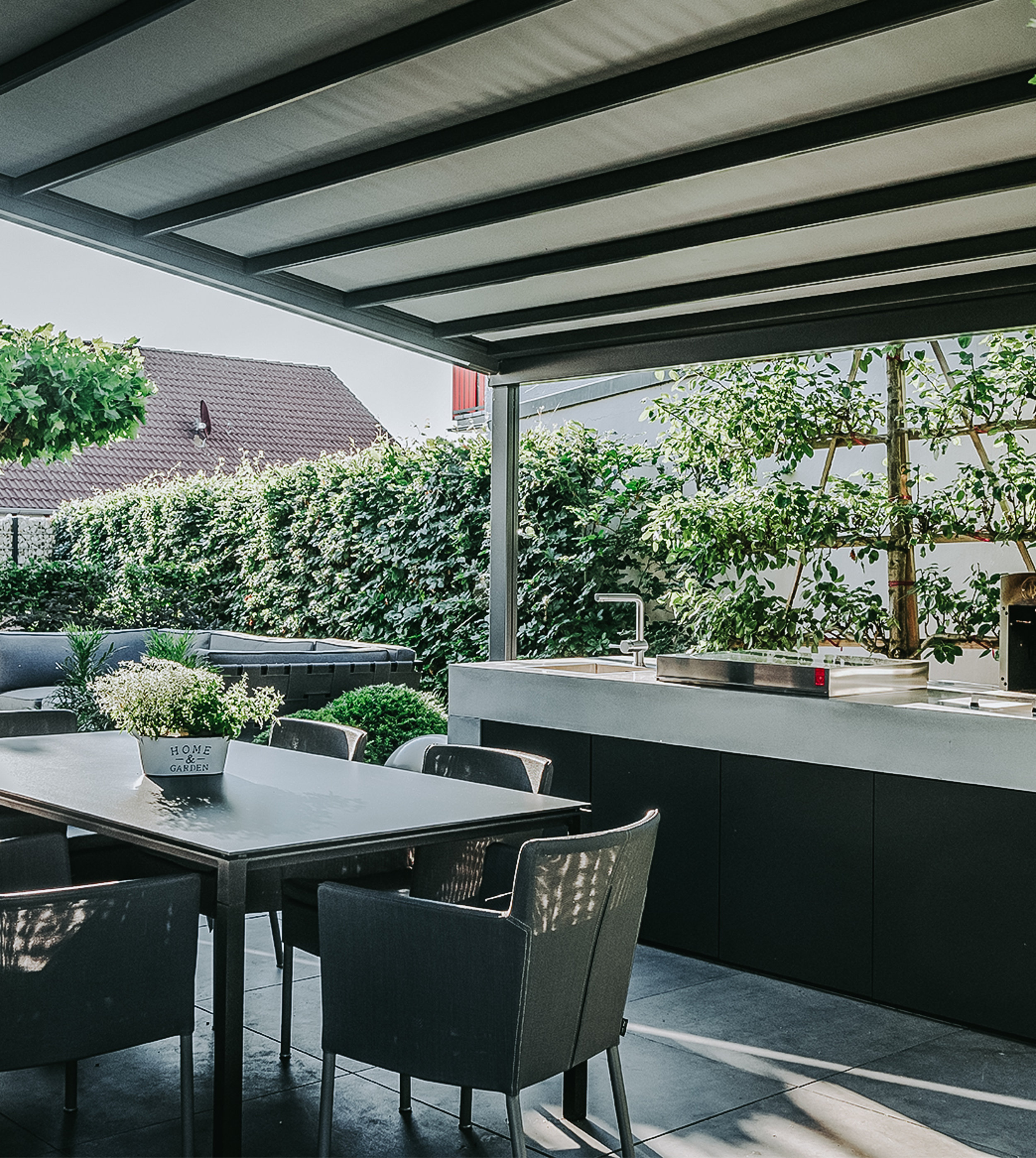 Eine überdachte Terrasse mit schwarzen Fliesen, einer Sitzgruppe und einer Gartenküche. Dahinter ist ein Outdoorsofa zu sehen sowie ein Sichtschutz aus Pflanzen.