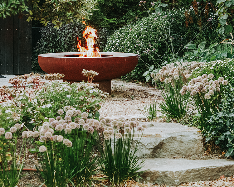 Rustikale Feuerschale im Garten umgeben von Steinen und klimaresilienten Pflanzen 