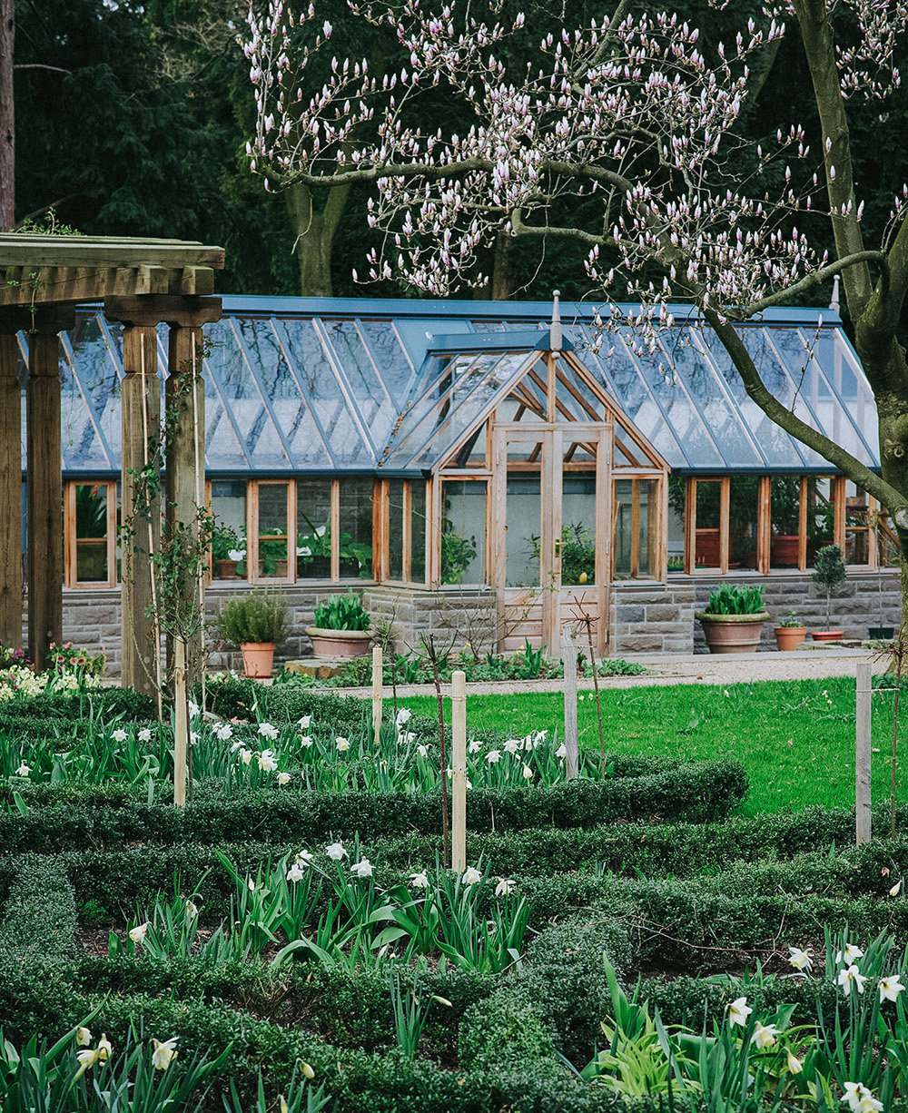 Gewächshaus aus Naturstein und Holz steht in einem Privatgarten mit mehreren Beeten und Rasenfläche