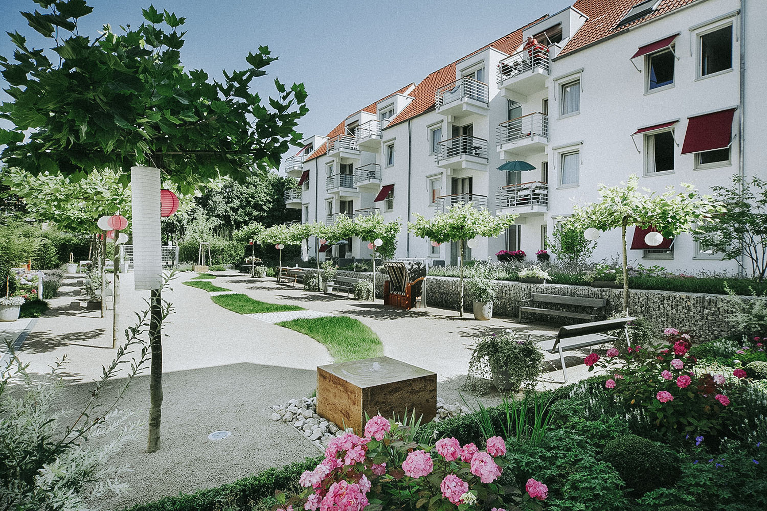 Innenhof aus Kies, in der Mitte ein Grünelement mit Brunnstein. Auf der Kiesfläche sind Gehölze gepflanzt und Sitzbänke angebracht.