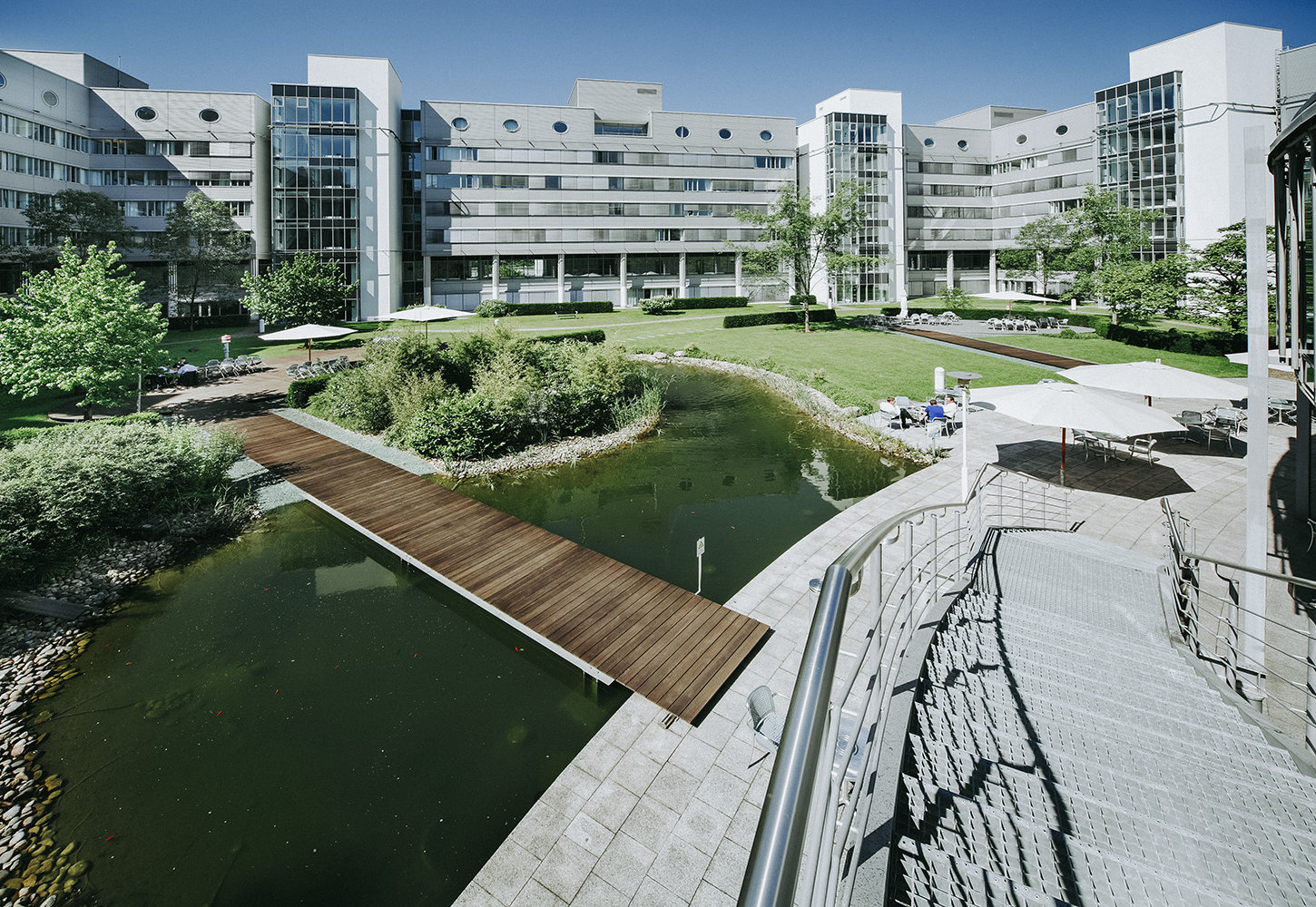 Firmengarten aus Vogelperspektive mit Treppe, einem großen Teich über den eine Holzbrücke führt sowie eine Terrasse mit Sonnenschirmen und eine grüne Rasenfläche. Der Garten befindet sich in der Mitte eines großen Bürokomplexes.