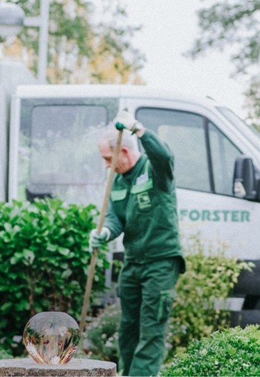 Ein Forster-Mitarbeiter steht mit einer Schaufel in der Hand in einem Garten mit Beleuchtung und einem Auto von Forster im Hintergrund. 