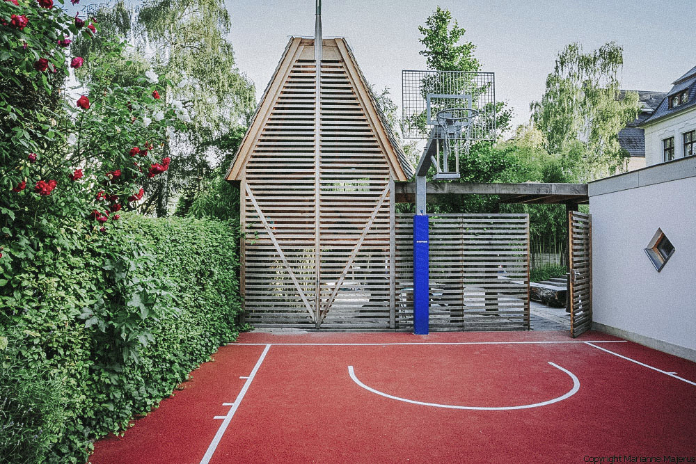 Ein Familiengarten in Koblenz mit Basketballfeld mit rotem Untergrund und Basketball Korb an einer Holzfassade. Auf der linken Seite ist eine grüne Sichtschutzhecke gepflanzt.