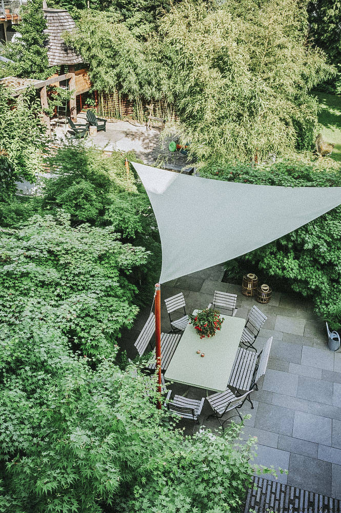 Ein Familiengarten in Koblenz mit einer Terrasse auf der eine Sitzgruppe steht. Die Terrasse wird von einem Sonnensegel überdach. Daneben befindet sich ein Spielbereich mit Baumhaus.