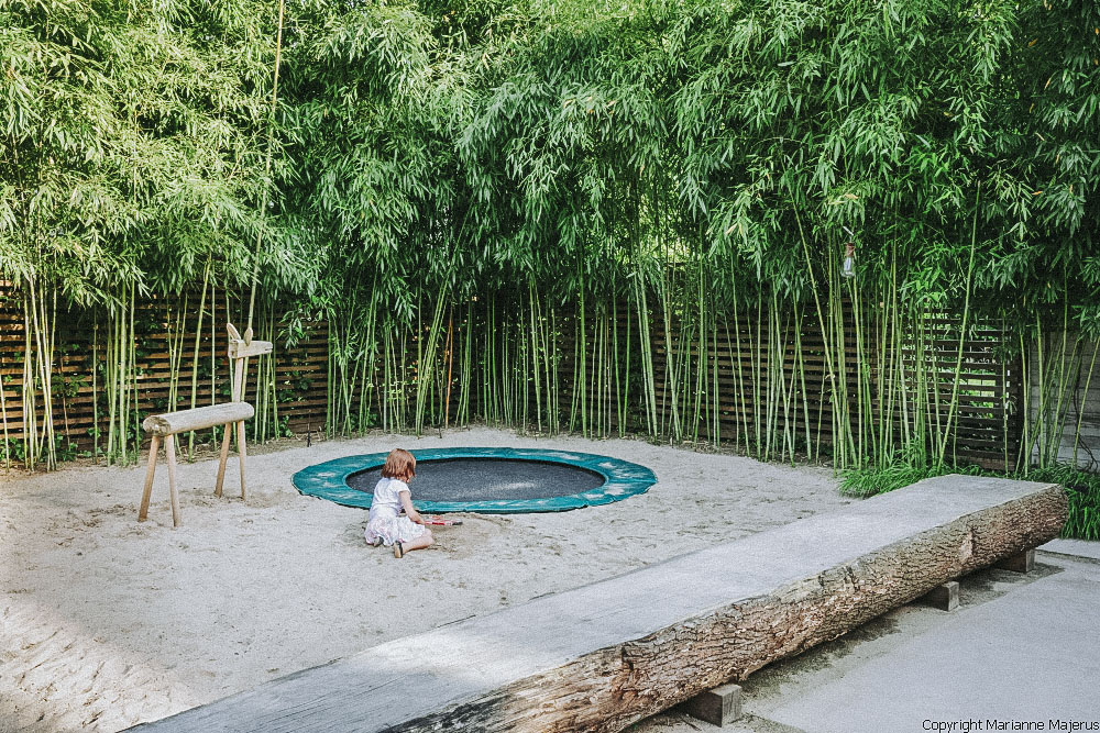 Ein Mädchen sitzt im Familiengarten in Koblenz im Sandkasten und spielt. Vor ihr ist ein Trampolin und ein Holzpferd. Um den Sandkasten ist Bambus gepflanzt. 