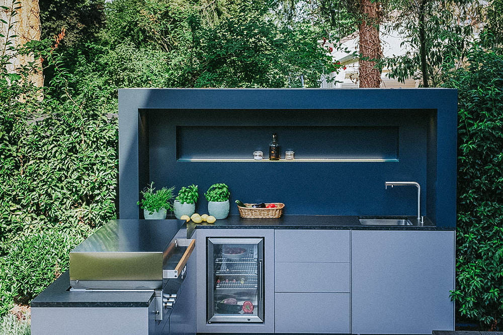 Eine graue hochwertige Gartenküche in Bonn mit eingebautem Grill, Kühlschrank und Waschbecken sowie einer installierten Beleuchtung