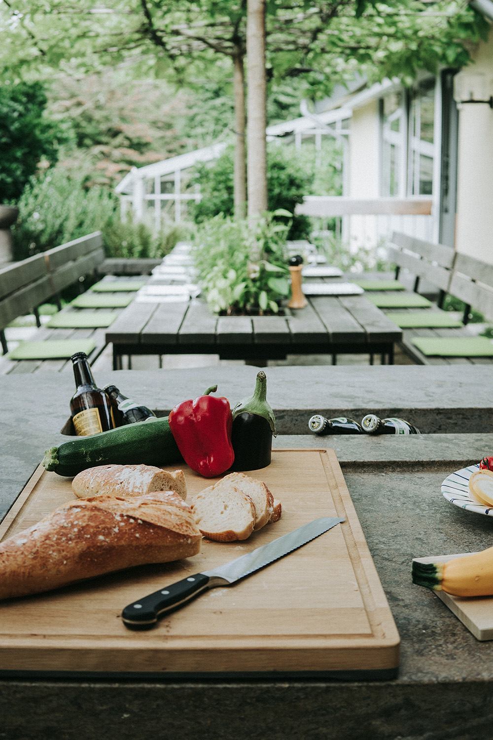 Auf der Arbeitsplatte aus Naturstein der Außenküche liegt ein Holzbrett mit Brot, Gemüse und einem Schneidemesser. Der Blick geht von der Arbeitsplatte in Richtung gedeckter Esstisch mit grüner Bepflanzung.