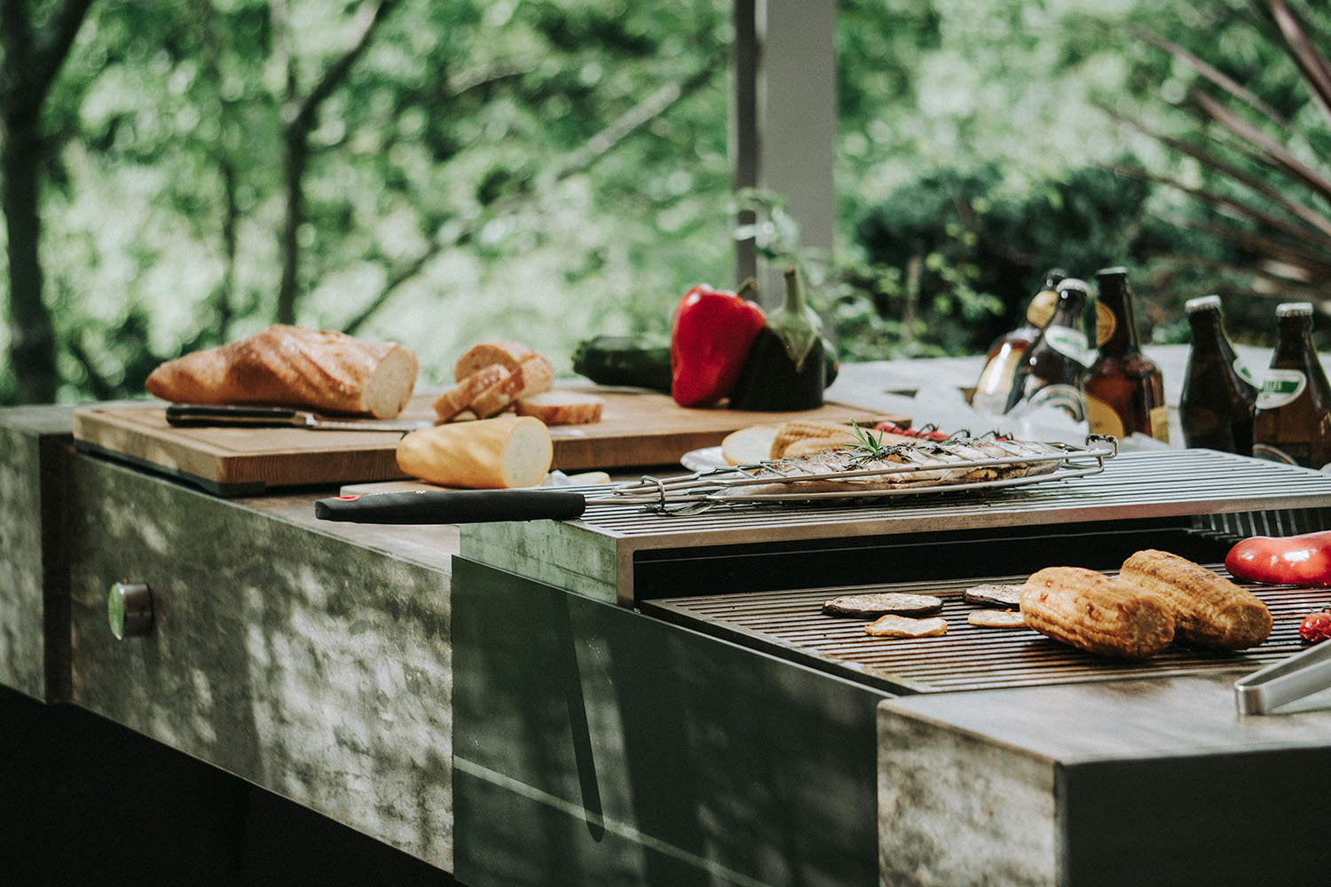 Eine Gartenküche aus Naturstein mit einer Grillplatte, auf der Fisch und Gemüse gegrillt werden. Daneben ein Holzbrett mit Brot.
