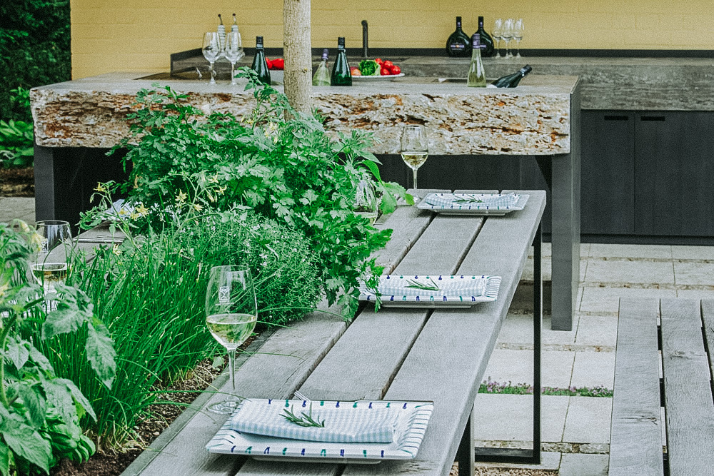 Ein gedeckter Gartentisch mit grüner Bepflanzung steht vor einer Gartenküche mit großer Natursteinplatte in Königswinter bei Bonn