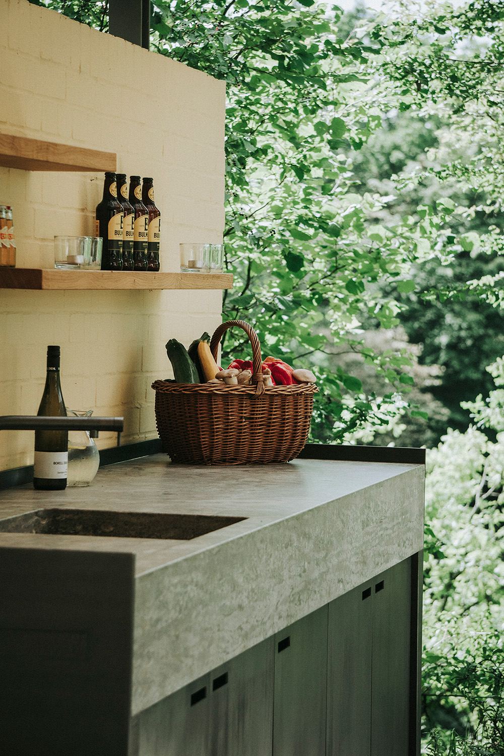 Eine Gartenküche aus Naturstein und Schwarzstahl. Der Küchenblock ist mit einem Wasserhahn und ausgestattet. Ein Korb mit Gemüse steht auf der Arbeitsplatte und Holregale hängen an der Wand. 