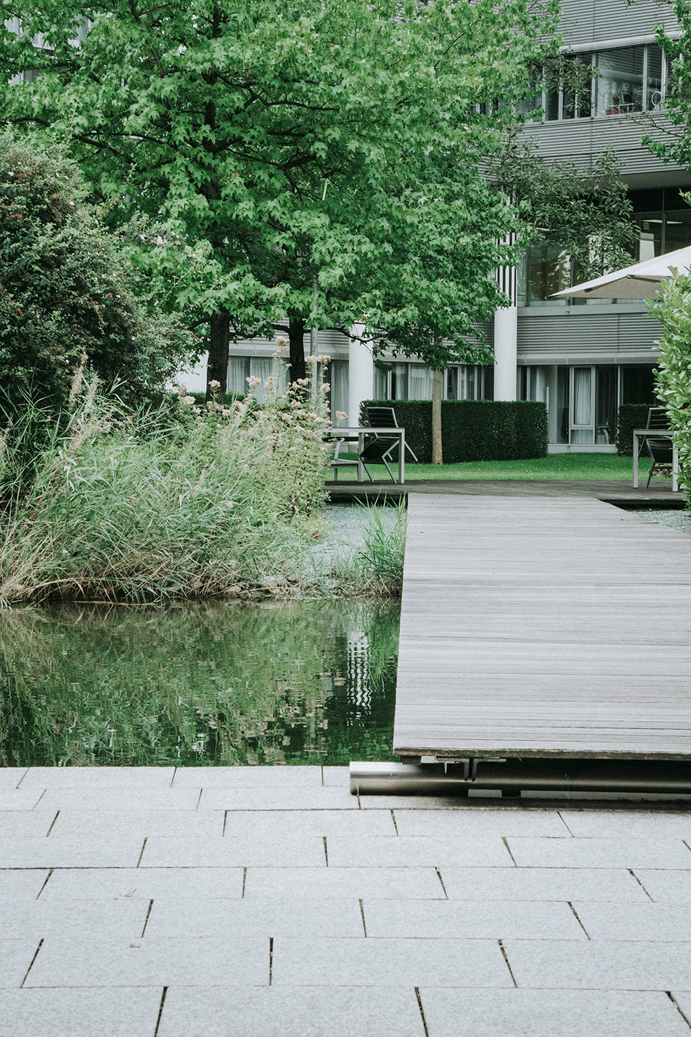 Eine Holzbrücke führt von einer grau gepflasterten Terrasse über einen Teich hinüber zu einem Sitzbereich mit Schirmen im grün angelegten Firmengarten.
