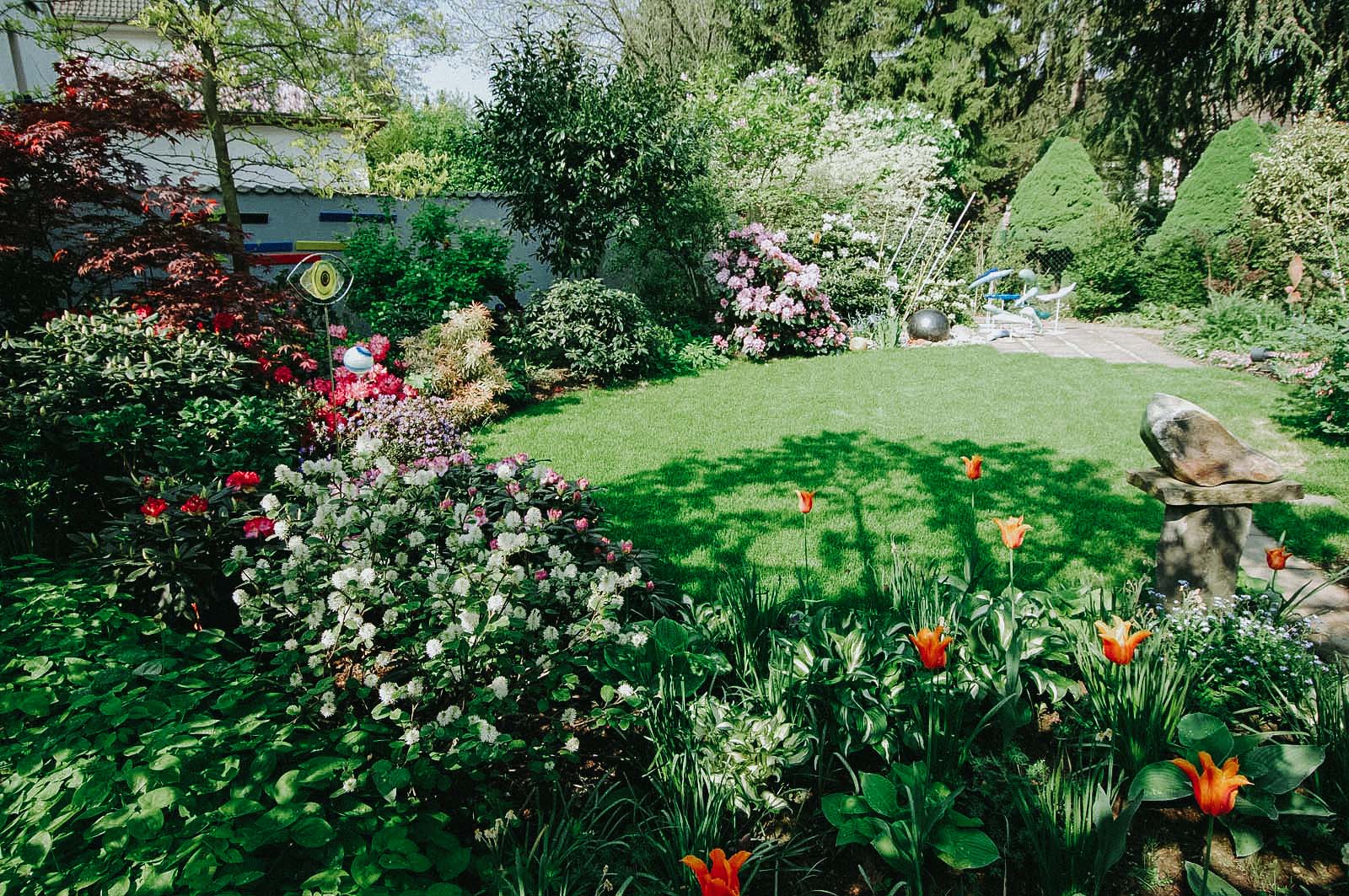 Ein kleiner Privatgarten mit runder Rasenfläche, dichter Staudenbepflanzung mit Deko-Elementen, Formgehölzen sowie Kunstobjekte