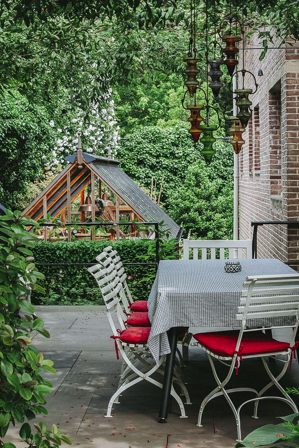 Eine grün bepflanzte Terrasse mit Sitzbereich aus Tisch und Stühlen. Der Blick geht auf das Gewächshaus aus Holz und Glas