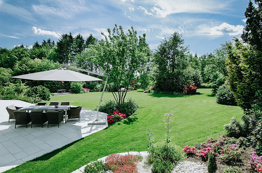 Privatgarten mit Terrasse, Gartenmobiliar, Ampelsonnenschirm sowie eine Rasenfläche mit mehreren Staudenbeeten und Gehölzen.